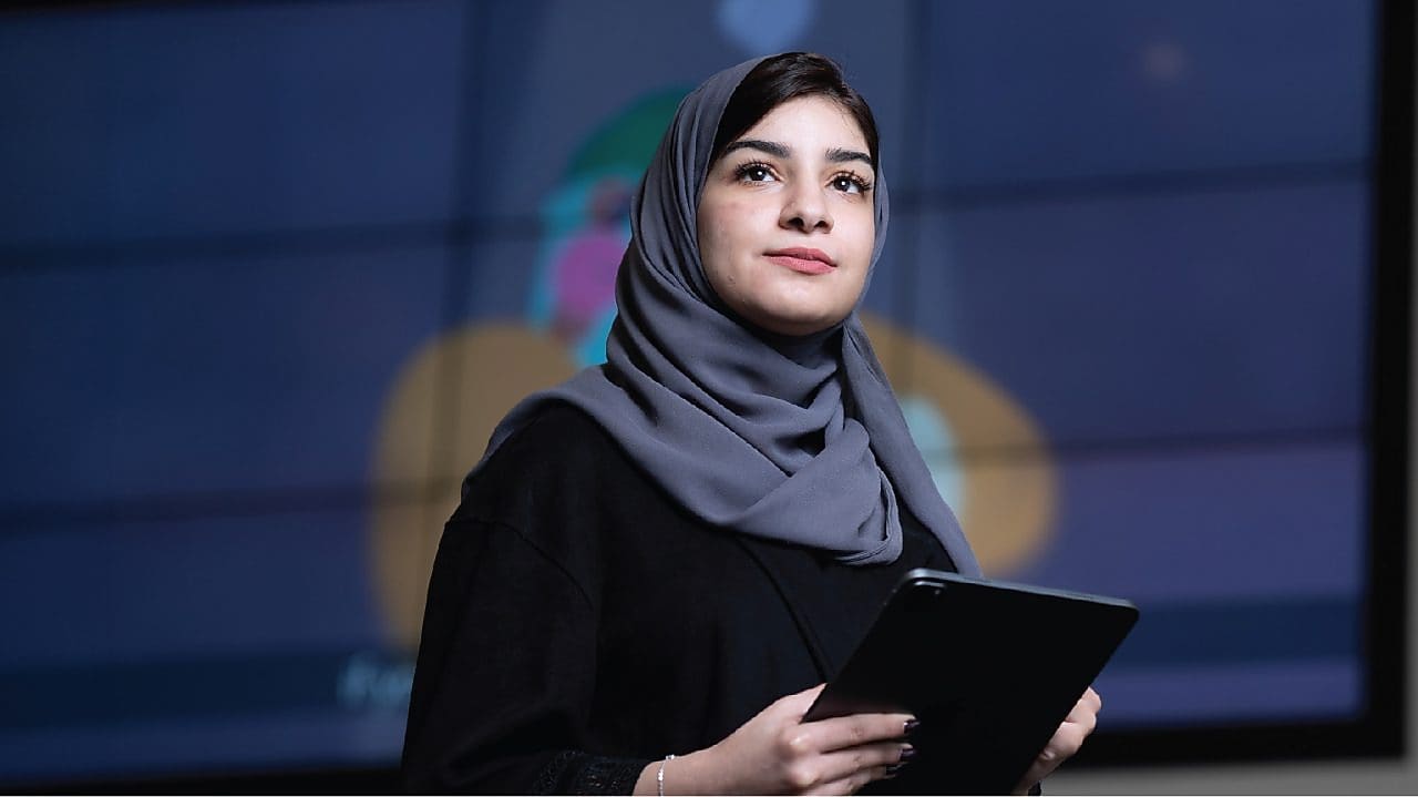 A person wearing a grey headscarf and black clothing is holding a tablet