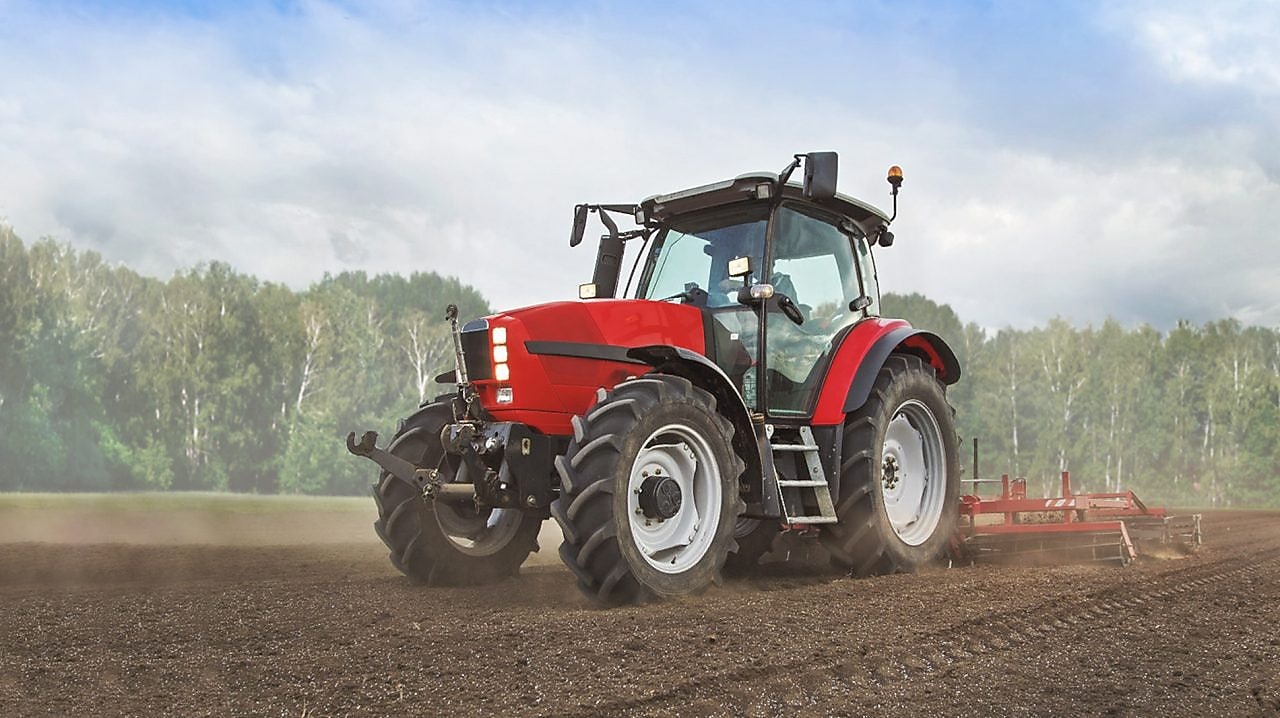 A red tractor ploughing a muddy field with trees in the background