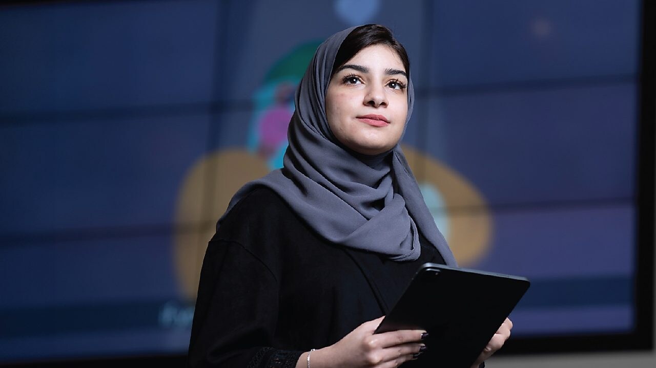 A person wearing a grey headscarf and black clothing is holding a tablet
