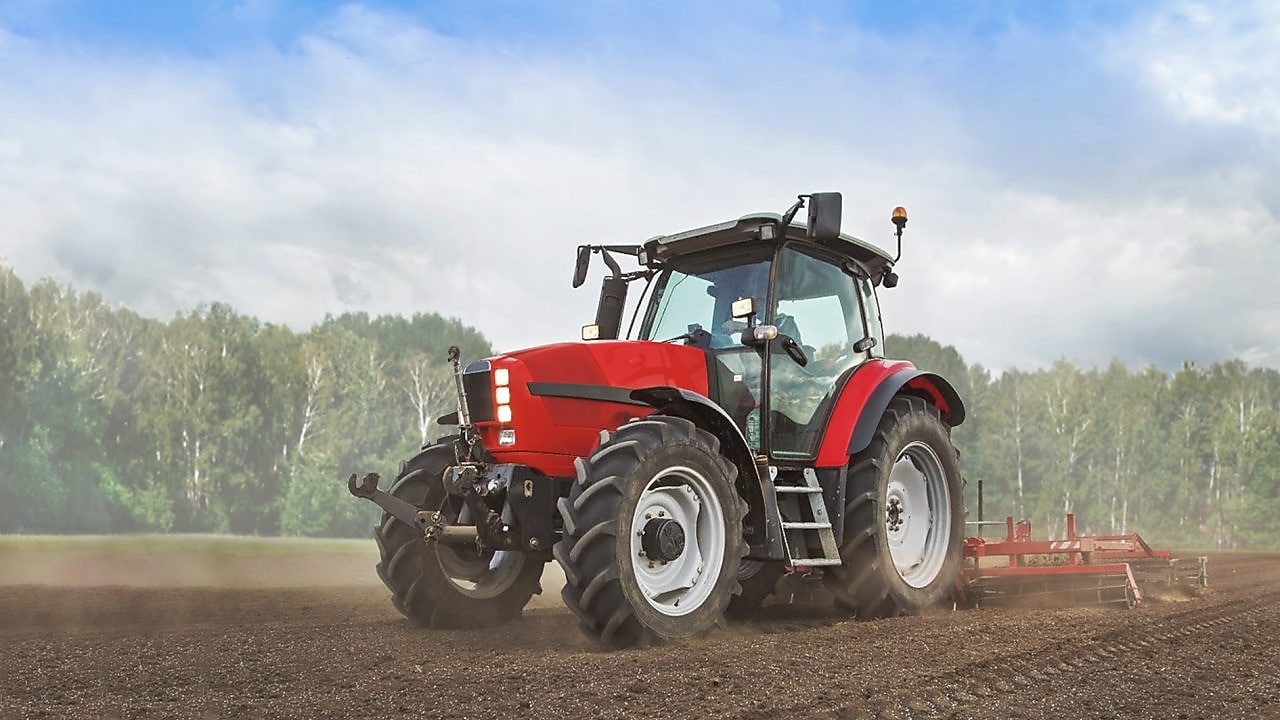 A red tractor ploughing a muddy field with trees in the background
